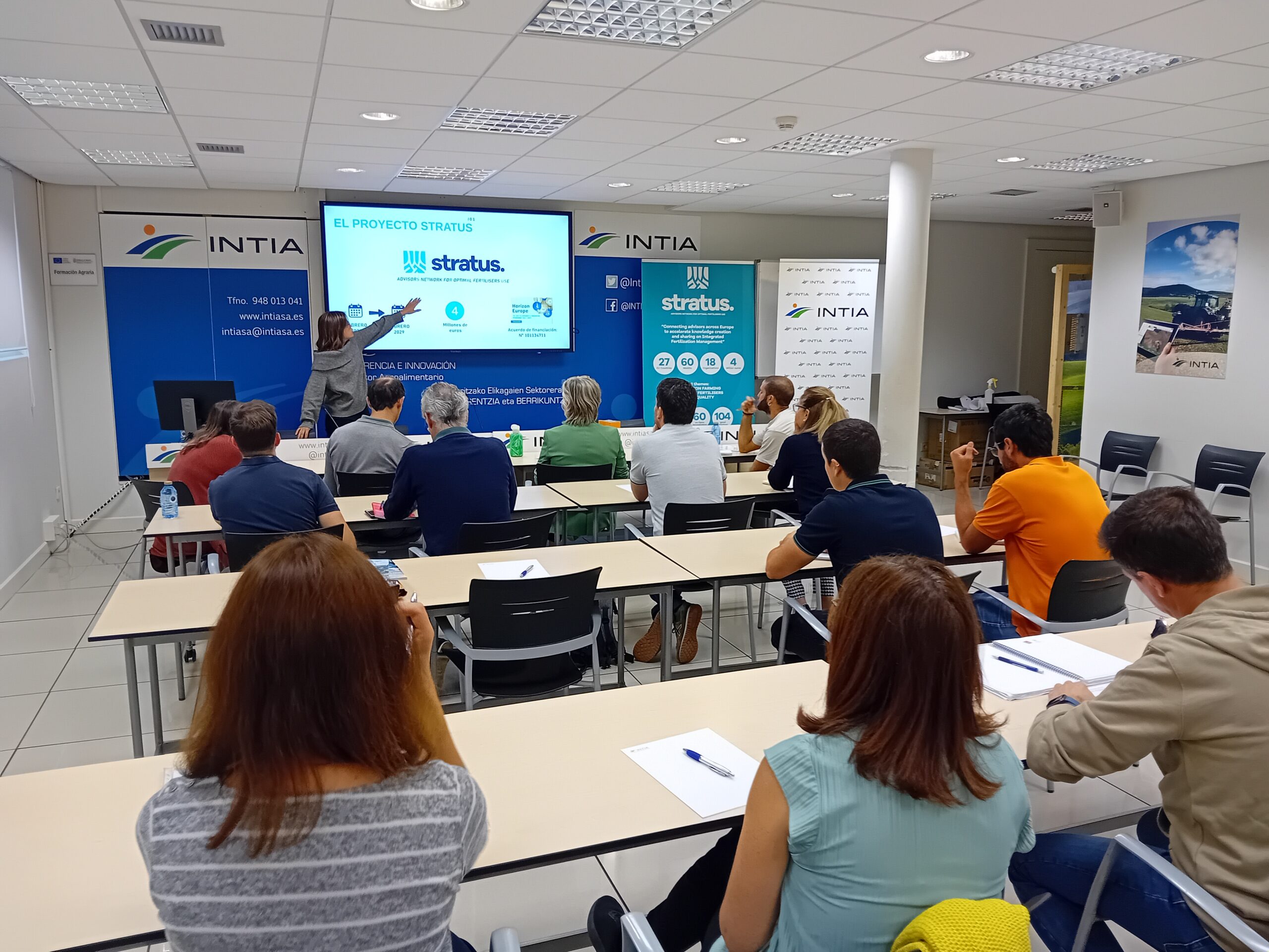 The image shows a group of people attending a presentation in a classroom setting. The presenter, standing at the front, is pointing to a screen displaying a slide about the STRATUS project, which includes key statistics and objectives. The room is set up with tables, and participants are seated, taking notes or listening attentively. The setting appears professional, with branding from INTIA and STRATUS visible on banners and walls, emphasizing the focus on agricultural innovation and sustainable fertilization practices.