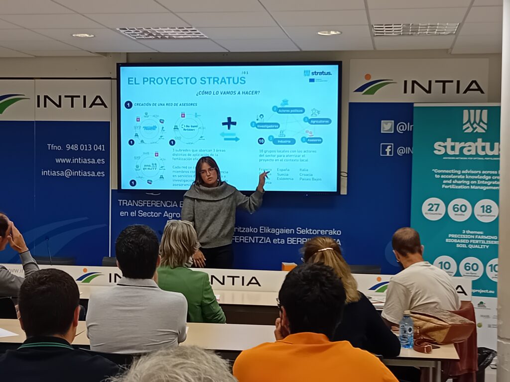 A person is giving a presentation in front of a room. The presenter is pointing to a projection screen showing various texts and graphics related to 'THE STRATUS PROJECT'. The audience, consisting of several people, is looking at the presenter and the screen. On the screen are diagrams, bullet points and icons of clouds, people and technological devices. There are logos in the lower right corner of the screen that appear to be associated with the project or the organizations involved.