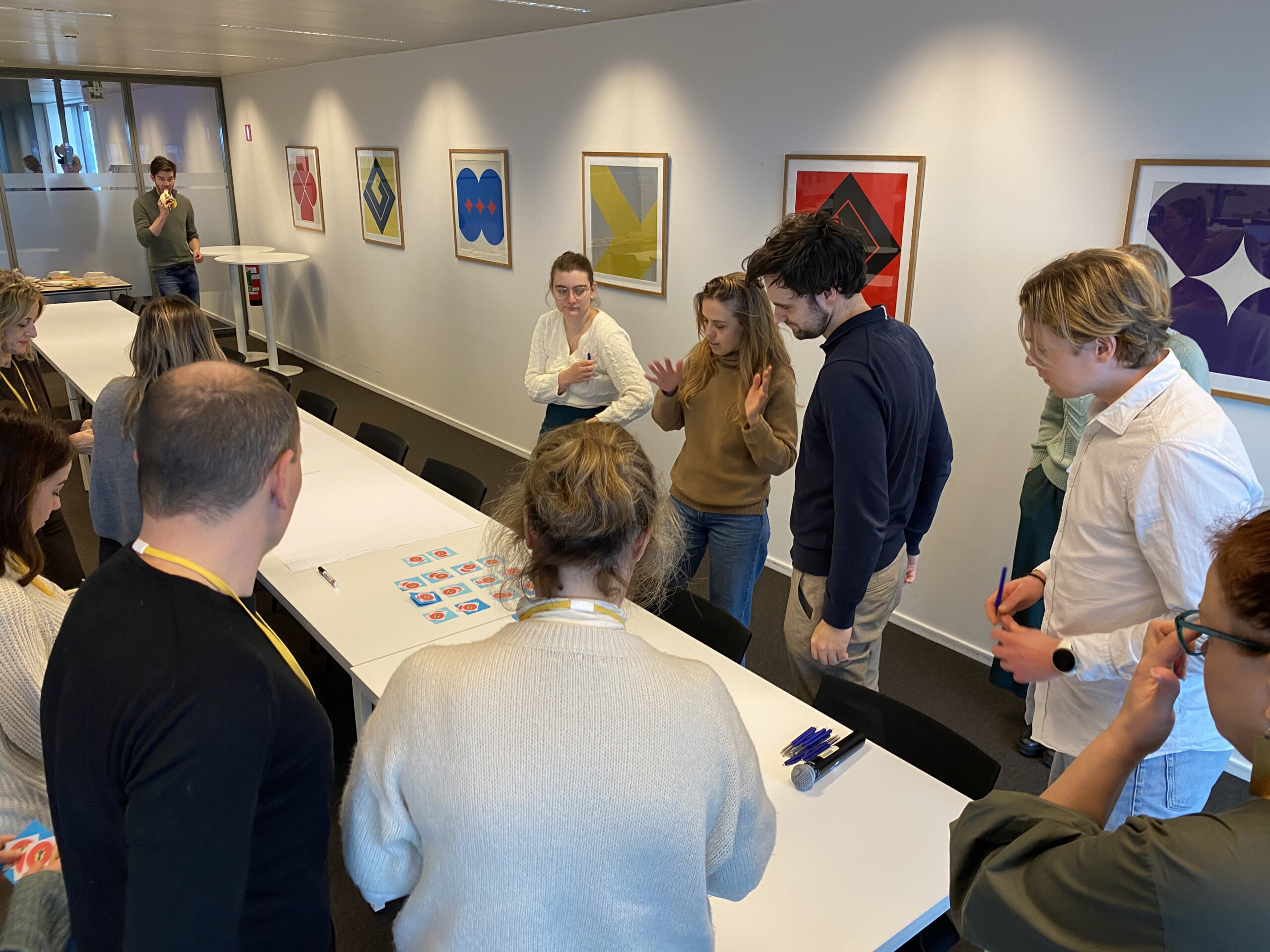 A group of people stands around a long table in a meeting room, collaborating on an activity with printed cards. Several framed abstract artworks hang on the wall, and another person speaks from a podium in the background.