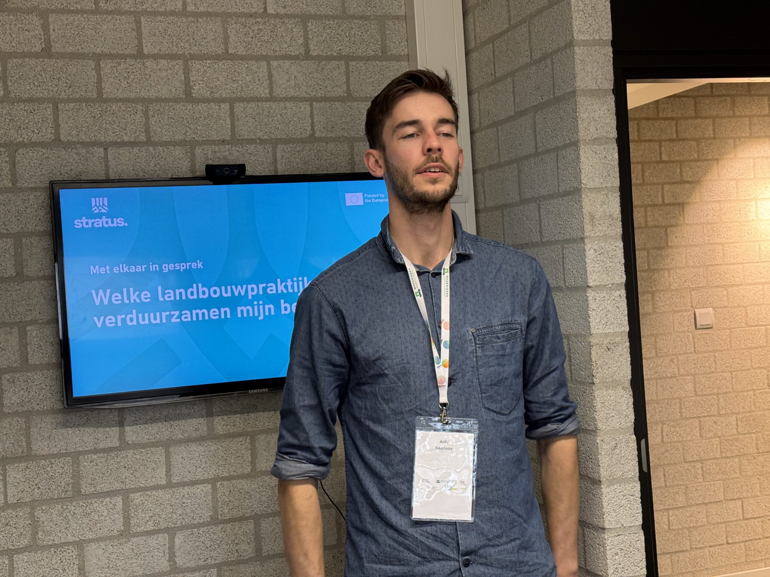 A participant standing in front of a presentation screen displaying information about sustainable agricultural practices during a Stratius event, wearing a lanyard and badge in a conference setting.
