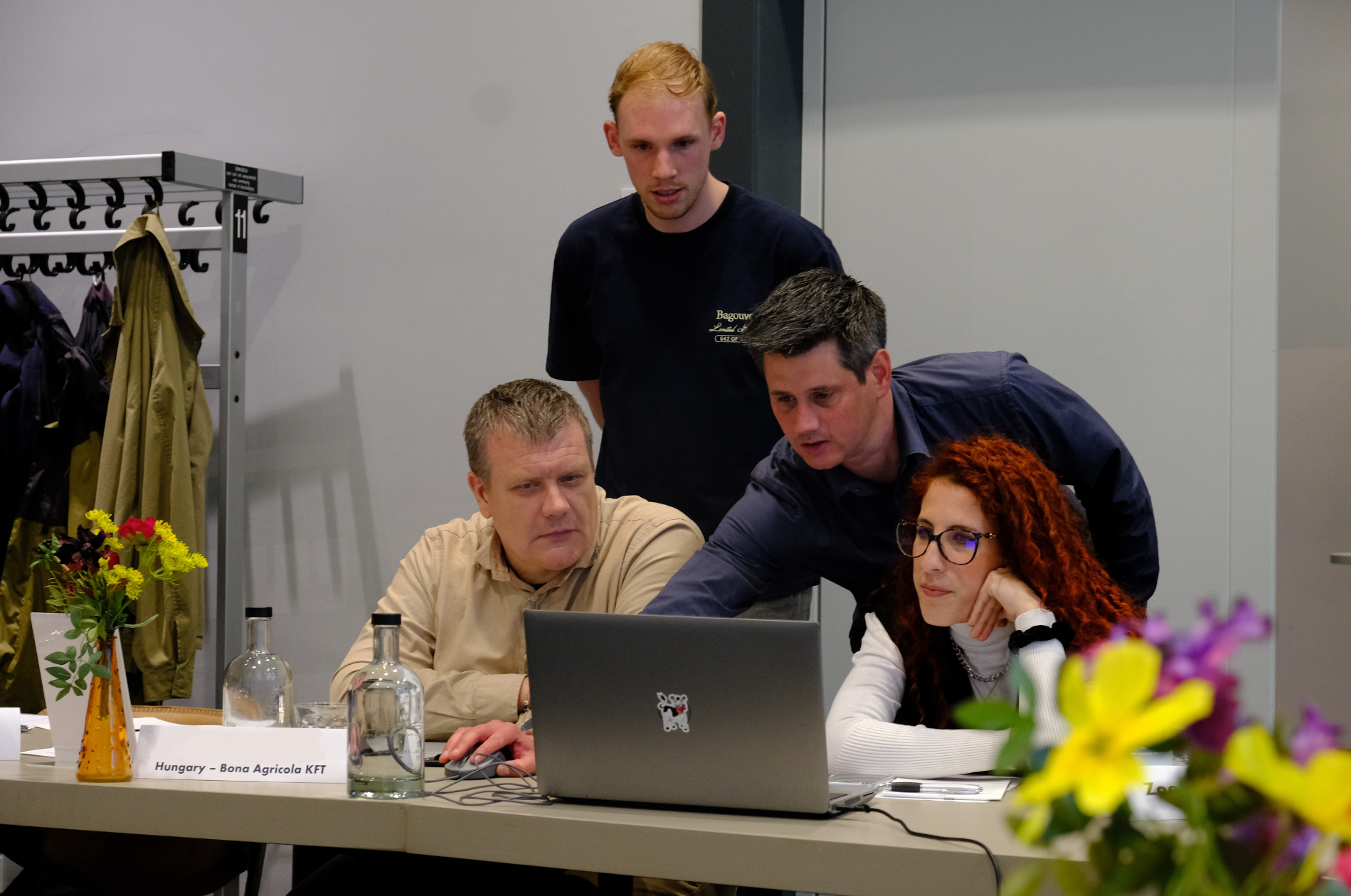 Four people collaborate around a laptop at a conference or meeting table. One seated person operates the laptop while another leans over to point at the screen. A third seated participant and a fourth standing participant look on. The table contains water bottles, papers, name placards, and flower arrangements, with a coat rack visible in the background.