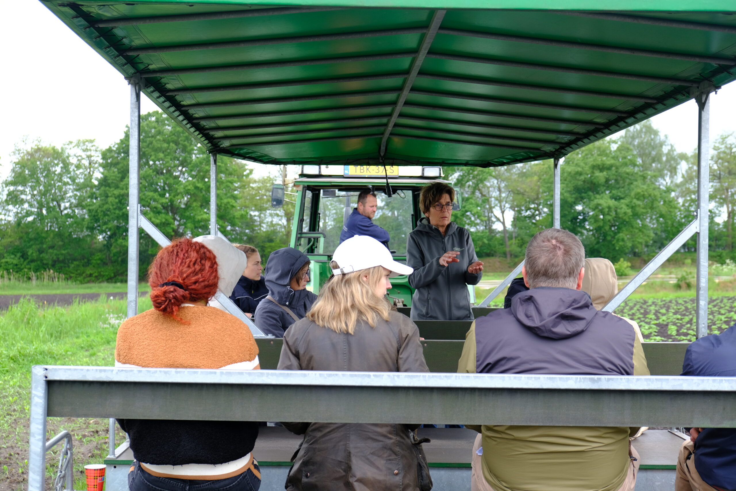 A group of people sit on a covered farm wagon while an instructor stands at the front, speaking to the group. The wagon is parked in an agricultural field with rows of crops and trees visible in the background, and a tractor is attached to the wagon.